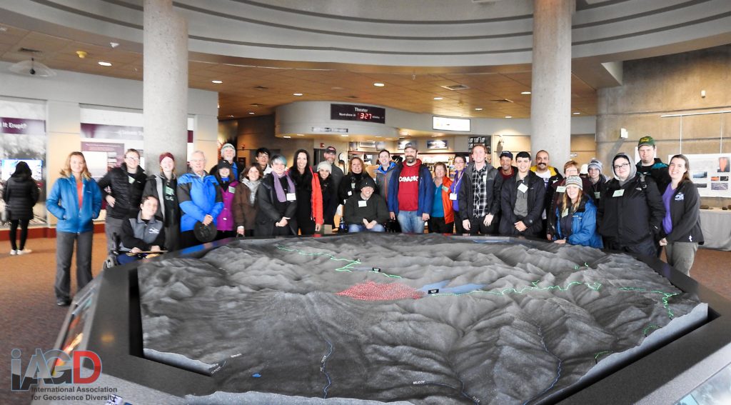 A group of 30 people stand and sit behind a large indoor 3D model of Mount Saint Helens. 