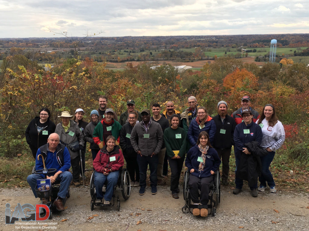 21 people stand or sit in wheelchairs in front of fall foliage and an overlook of a valley.