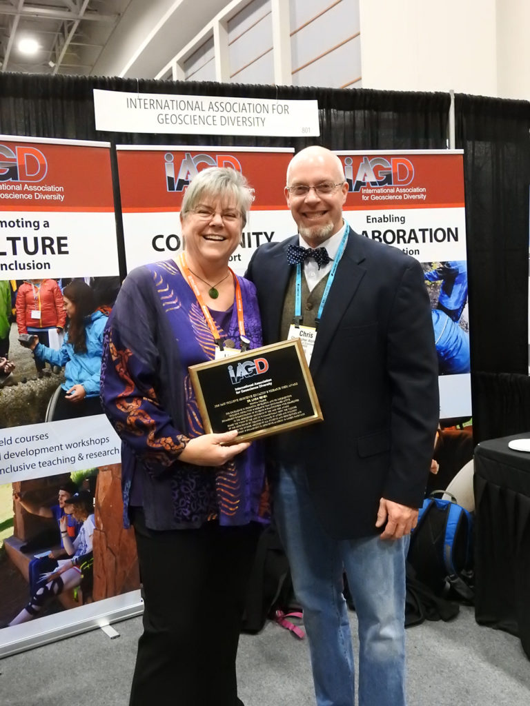 Linda Reinen and Chris Atchison stand in front of the IAGD booth holding a plaque