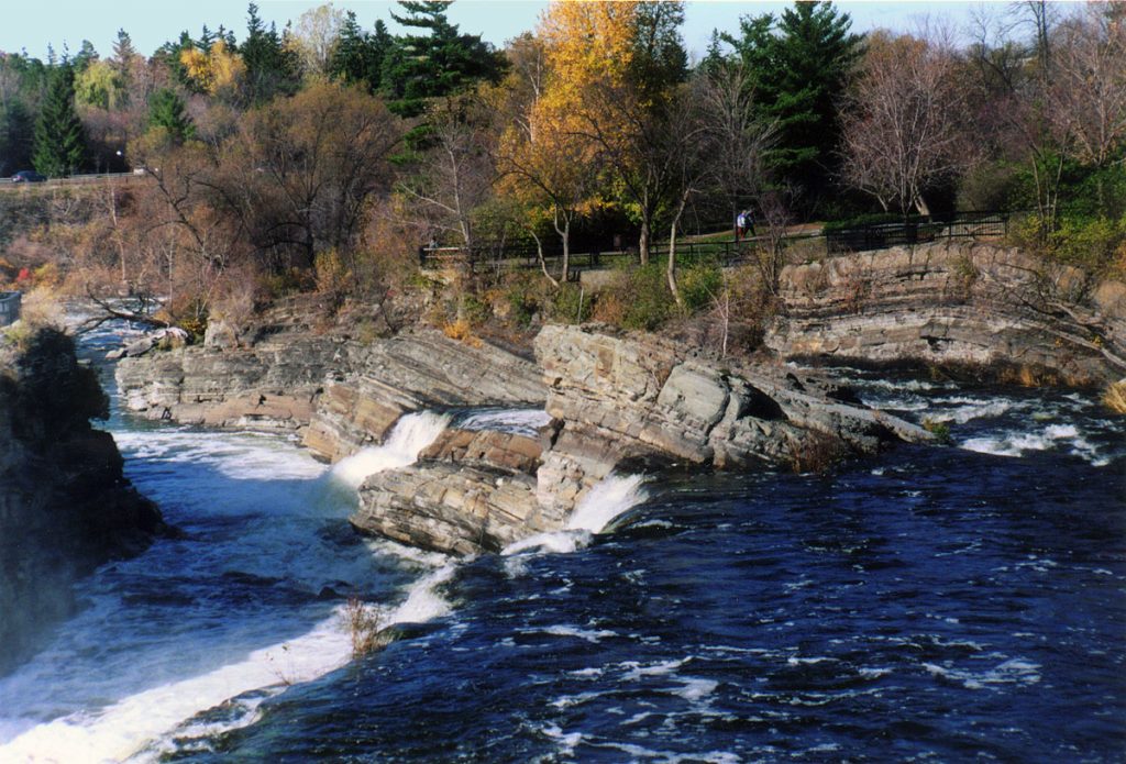 Leaves in Fall colors and bright blue water frame rock out crops of tilted sedimentary rock.