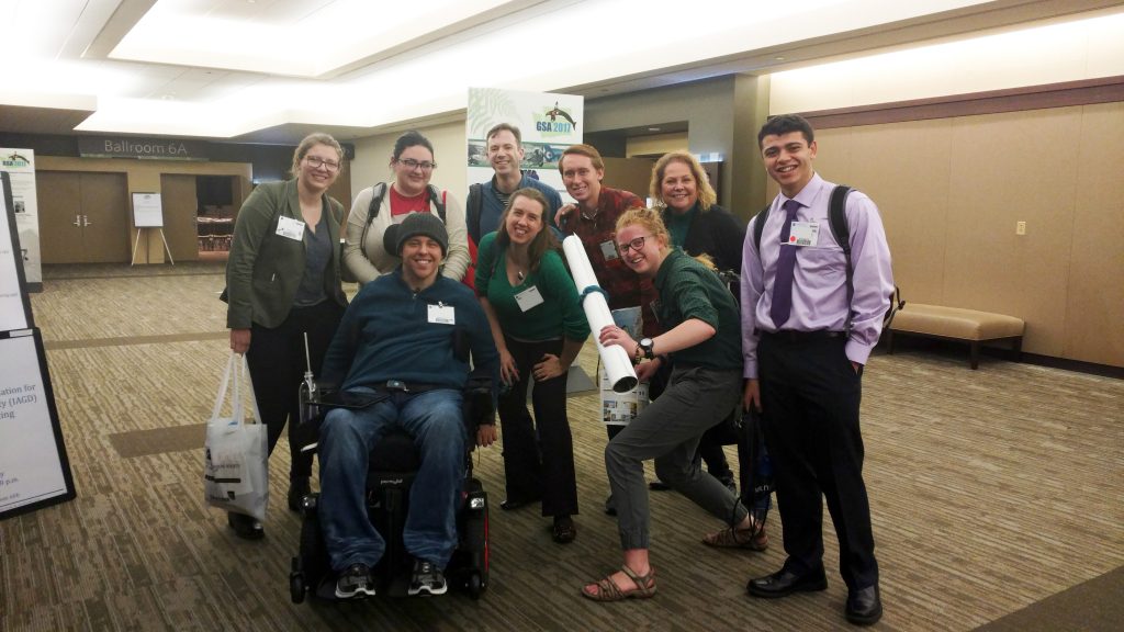 A group of students and faculty stand and sit in front of a banner advertising GSA 2017 in Seattle.