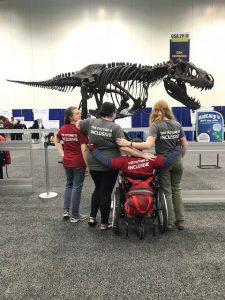 Four people (three standing and one in a wheelchair) in front of a T-Rex skeleton. All four have red and gray shirts that read "The Future is Inclusive" on the back.