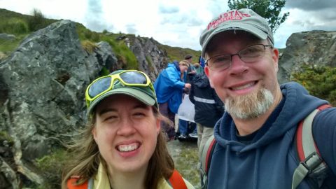 Anita and Chris smile for a selfie. Both are wearing ball caps, backpacks and cool weather gear. Students are working on documenting a rock outcrop in the background.