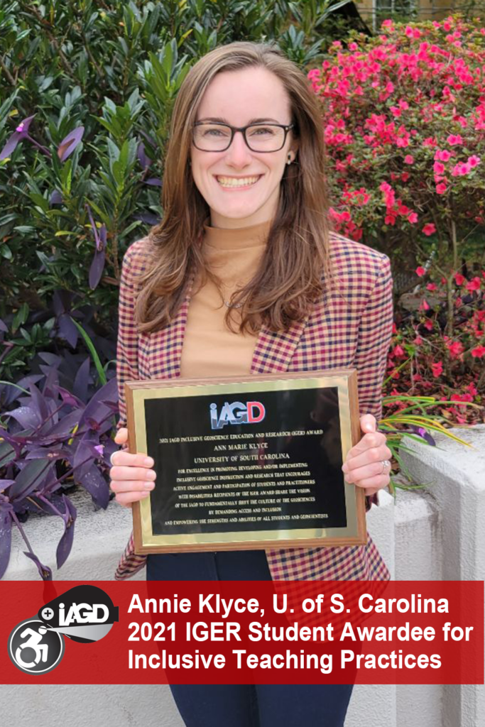 Annie Klyce, smiles and holds her IGER award plaque in front of colorful flowering plants.