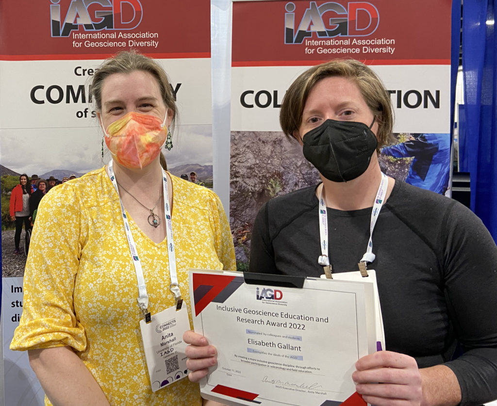 Lis Gallant holds a certificate in front of IAGD banners. Anita Marshall stands next to her. Both are wearing conference badges and masks.