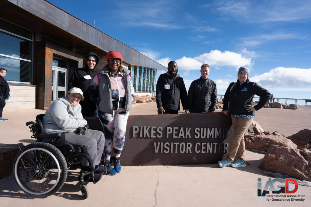 A group 6 people sit and stand around a sign that says Pikes Peak Visitor Center under bright blue sky with fluffy clouds that look quite low due to the altitude.