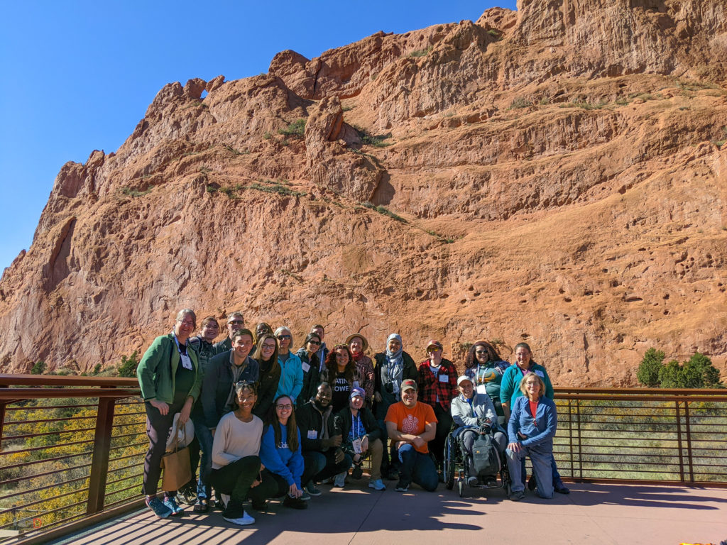 Group photo of a diverse group of students and faculty in front of the massive red rock formation known as the "Kissing Camels".