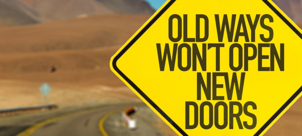 A yellow road sign on a winding desert road says "Old ways won't open new doors"