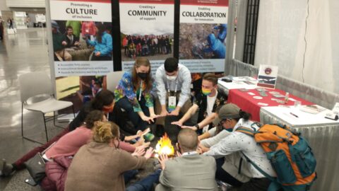 visitors to the IAGD booth at GSA conference huddle around a cardboard fire and pretend to warm their hands.