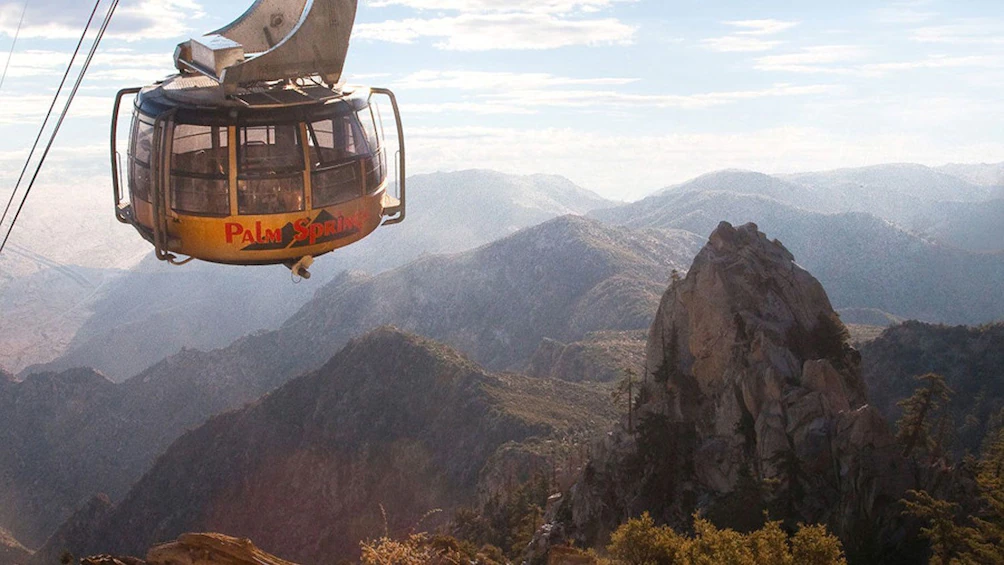 A large round glass enclosed gondola hangs from a cable with dramatic mountain views in the background