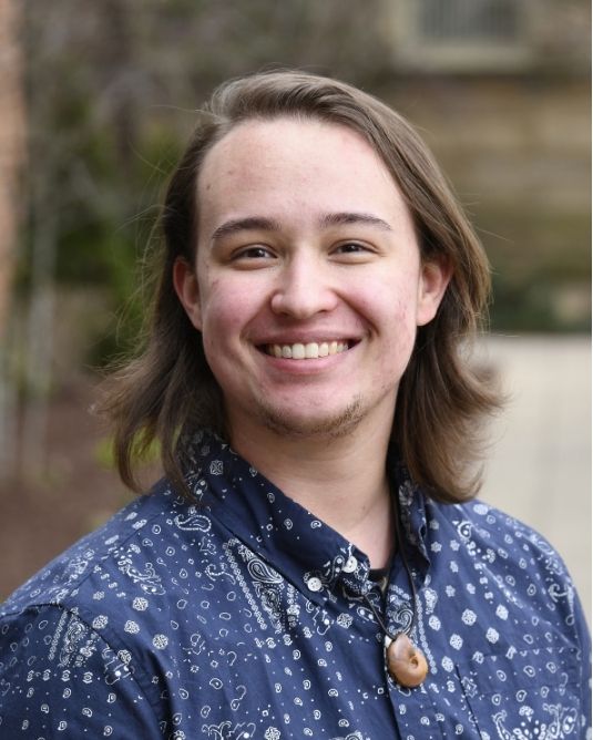 A smiling young caucasian individual with long wavy brown hair and incipient facial hair on the chin. The individual is wearing a dark blue collared shirt with white spotted patterns and a necklace affixed with a small light-brown ammonite fossil.The individual is outside set against a blurry background.
    