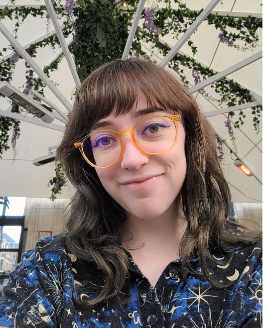 A young, white, female-appearing individual is shown smiling slightly. Frizzell has long slightly-wavy dark brown hair, is wearing glasses with light-orange coloured rim and wearing a astronomy-themed dark collored shirt. She is indoors with a bacckground of white roof with vine-type plants.
    